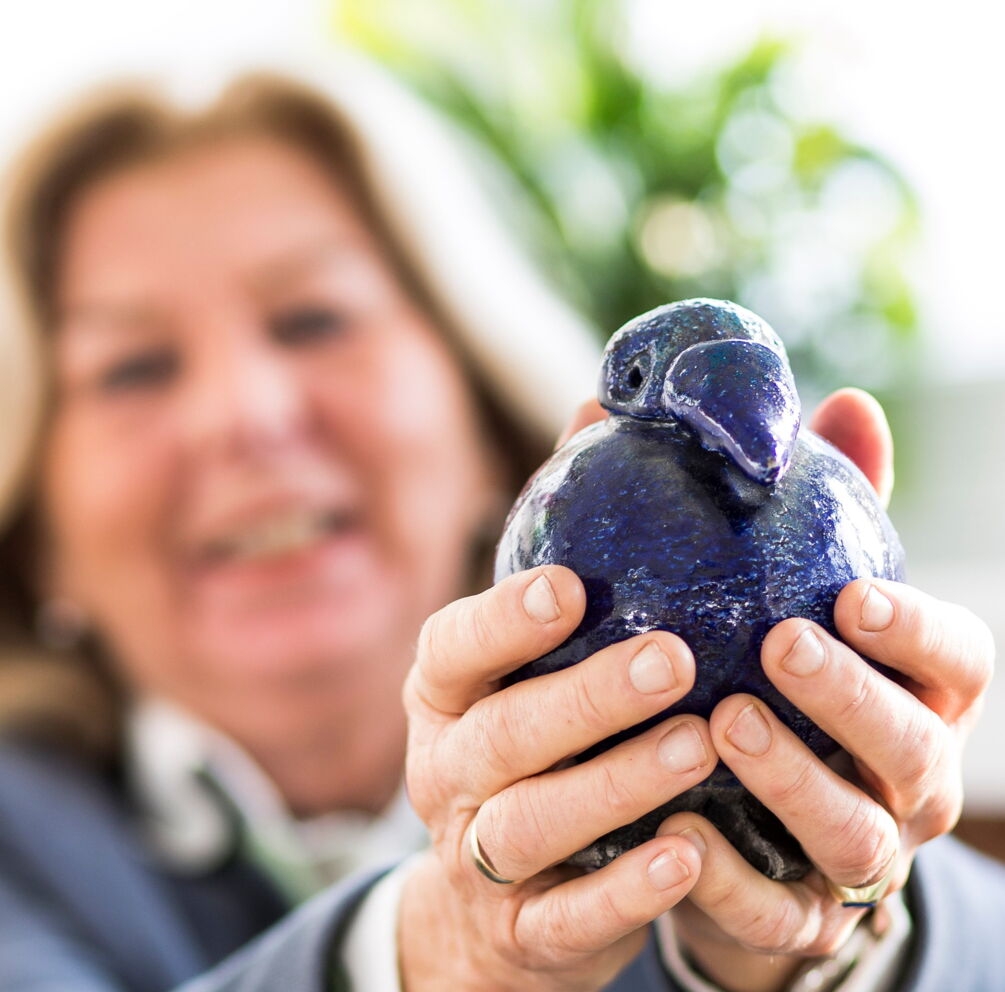 Eine Frau dessen Gesicht unscharf ist, während Sie Ihre Hände in die Kamera streckt und eine blaue Vogel Vase hält.