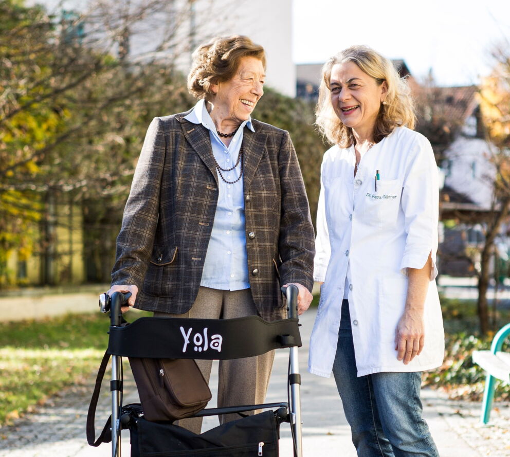 Frau Dr. Petra Gürtner, wie Sie mit einer älteren Dame, die einen Rollator benötigt, draußen an der frischen Luft ist und miteinander lachen.
