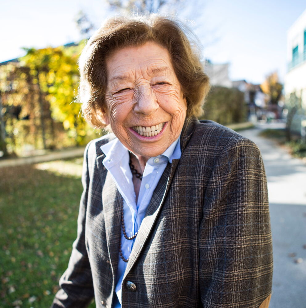 Ein Foto einer älteren Dame mit braunen Haaren und einem karierten Blazer, die lächelnd in die Kamera schaut.
