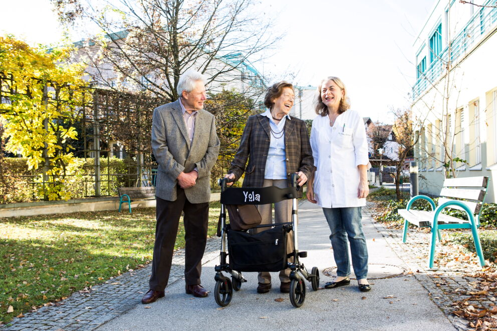 Zwei ältere Menschen, ein Mann und eine Frau die einen Rollator vor ihr hat, während sie mit Dr. Petra Gürtner lachen.