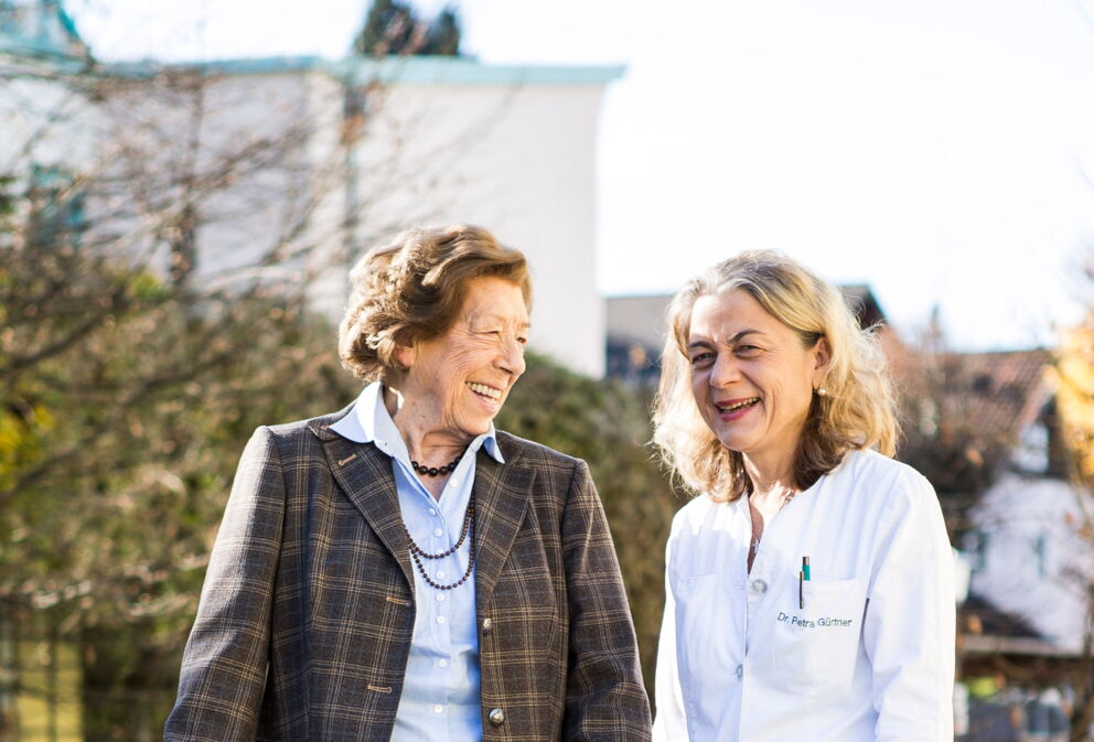Eine Patienten mit einem Rollator und Dr. Petra Gürtner die gemeinsam an der frischen Luft sind und lachen