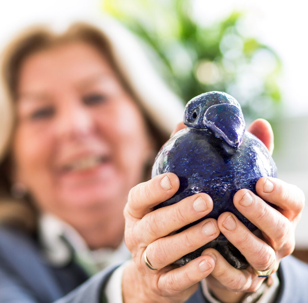 Eine Frau dessen Gesicht unscharf ist, während Sie Ihre Hände in die Kamera streckt und eine blaue Vogel Vase hält.